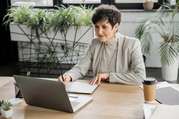 Досвідчена бізнес-леді, що пише в ноутбуці поблизу ноутбука і виймає напій, планування — стокове фото
