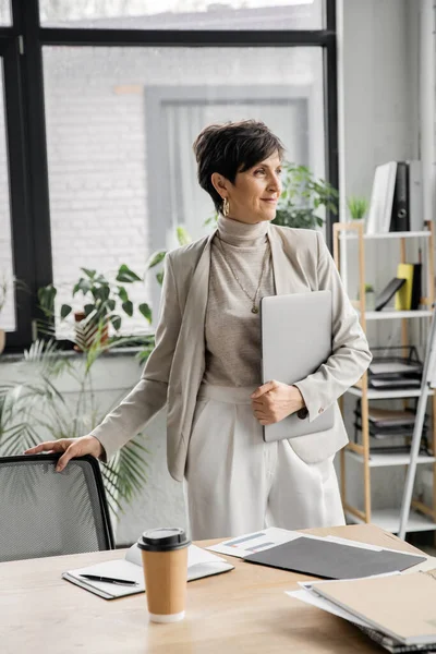 Positive Geschäftsfrau mittleren Alters mit Laptop, der in der Nähe von Dokumenten und Pappbechern wegschaut — Stockfoto