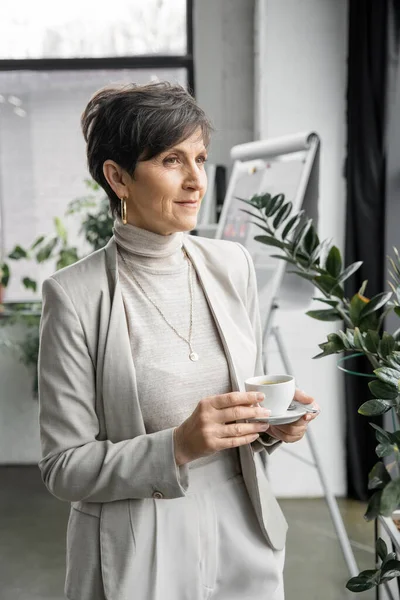 Confident and experienced middle aged entrepreneur with coffee cup looking away in modern office — Stock Photo