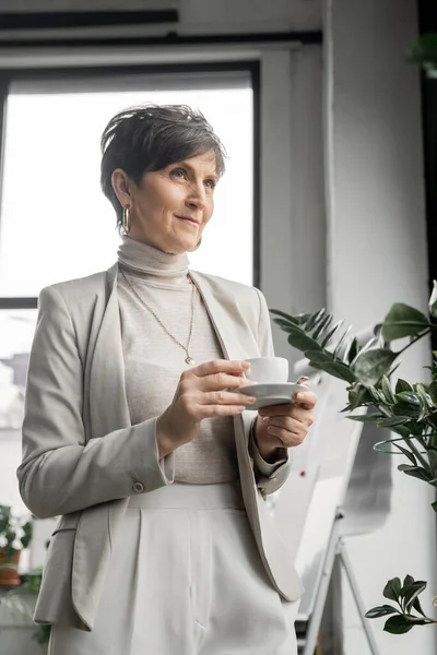 Femme d'affaires d'âge moyen positive et rêveuse avec tasse de café regardant loin dans le bureau moderne — Photo de stock