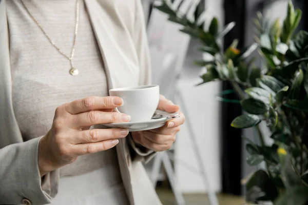 Vista ritagliata di elegante donna d'affari di mezza età in piedi con tazza di caffè in ufficio — Foto stock