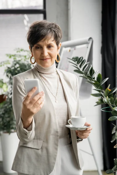 Positive middle aged businesswoman with coffee cup messaging on mobile phone in contemporary office — Stock Photo