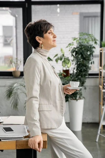 Reife Geschäftsfrau mit Kaffeetasse in der Nähe von Dokumenten und Smartphone im Büro, Seitenansicht — Stockfoto