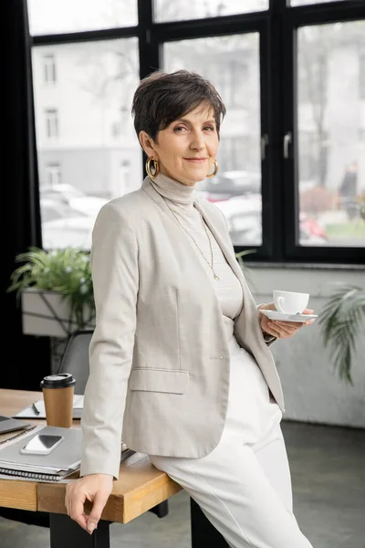 Successful middle aged businesswoman with coffee cup smiling at camera in modern office, headshot — Stock Photo