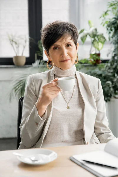 Professional headshot of successful businesswoman with coffee cup looking at camera near notebook — Stock Photo