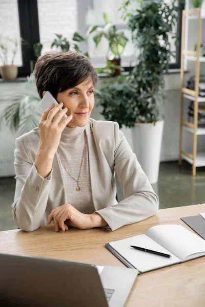 Успешный менеджер среднего возраста, разговаривающий на смартфоне рядом с ноутбуком и ноутбуком в офисе — стоковое фото