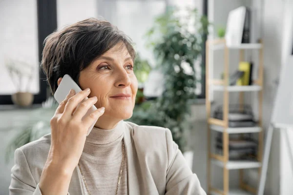 Elegante donna d'affari di mezza età che parla su smartphone e distoglie lo sguardo in ufficio moderno — Foto stock
