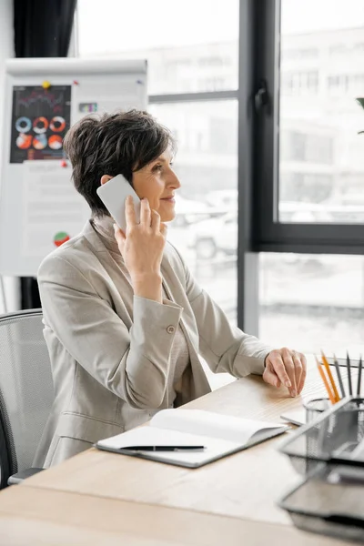 Оптимістичний менеджер середнього віку розмовляє на мобільному телефоні біля блокнота на робочому місці в сучасному офісі — стокове фото