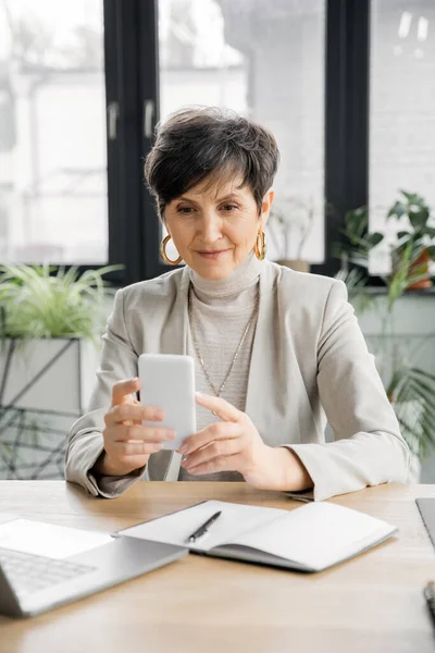Elegante mujer de negocios de mediana edad en red en el teléfono inteligente cerca de la computadora portátil y portátil en la oficina moderna - foto de stock