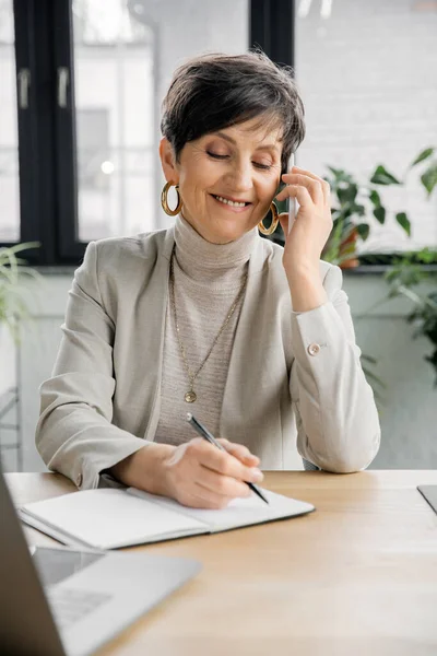 Lächelnde Geschäftsfrau mittleren Alters spricht auf dem Smartphone und schreibt in einem modernen Büro in ein Notizbuch — Stockfoto