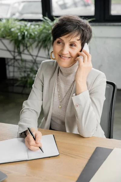 Positive mature corporate manager talking on smartphone, writing in notebook at workplace in office — Stock Photo