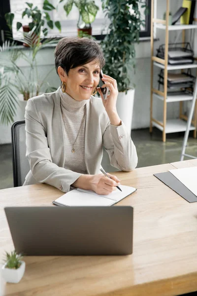 Pleased middle aged entrepreneur writing in notebook while talking on mobile phone in office — Stock Photo