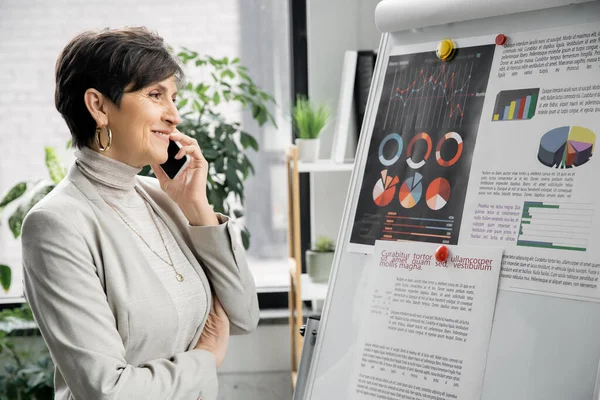 Радостная зрелая деловая женщина разговаривает на смартфоне возле флип-чарта с аналитикой в офисе, планируя — стоковое фото