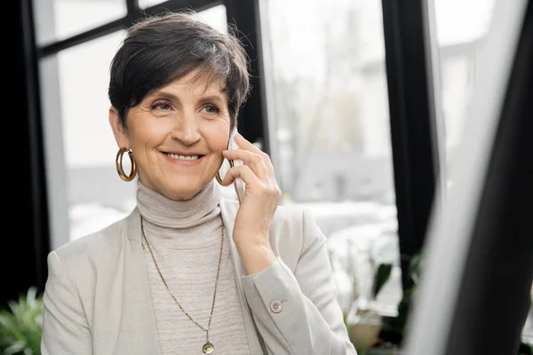 Empreendedor de meia-idade sorrindo e falando no smartphone no escritório moderno, sucesso empresarial — Fotografia de Stock