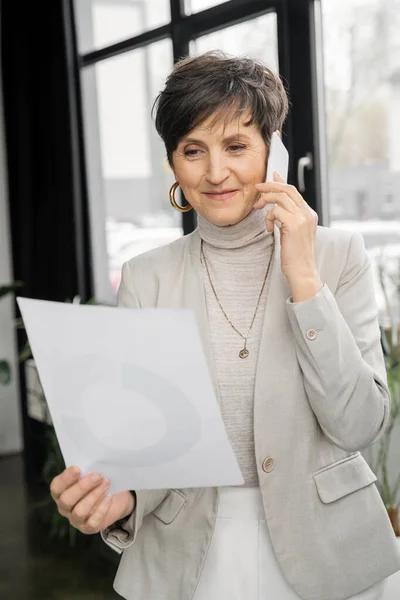 Mujer de negocios satisfecho mirando documento mientras habla en el teléfono móvil en la oficina contemporánea - foto de stock
