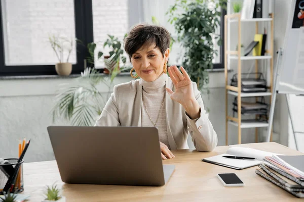 Sonriente mujer de negocios de mediana edad, saludando la mano, chat de vídeo en el ordenador portátil cerca de teléfonos inteligentes y portátiles - foto de stock