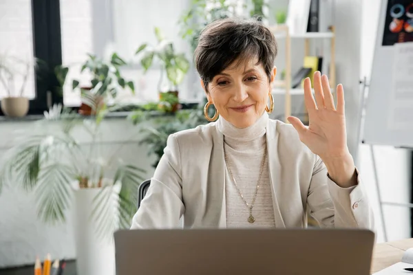 Donna d'affari matura positiva che agita la mano durante videoconferenza su computer portatile in ufficio moderno — Foto stock