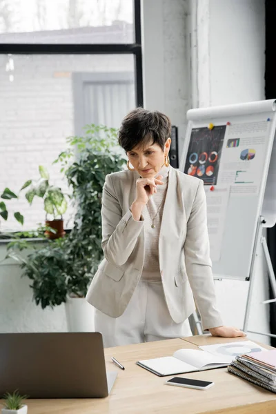 Thoughtful mature entrepreneur near laptop and flip chart with graphs, video chat, problem solving — Stock Photo