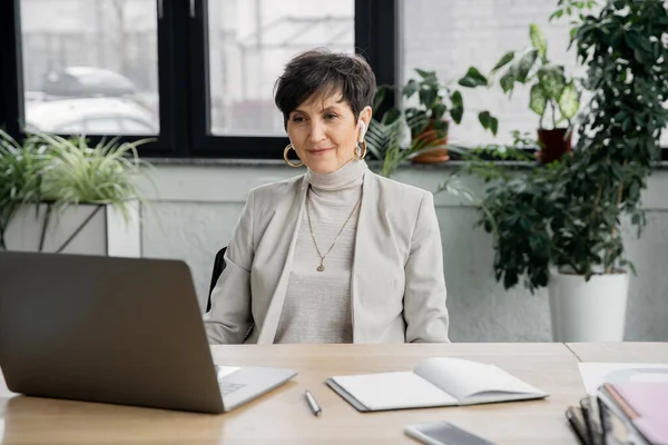 Donna d'affari matura che guarda laptop vicino a notebook e smartphone sulla scrivania in ufficio — Foto stock