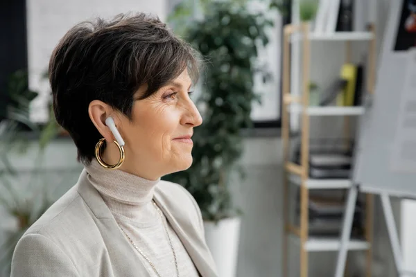 Side view of cheerful middle aged businesswoman in earphone listening music in office, enjoyment — Stock Photo