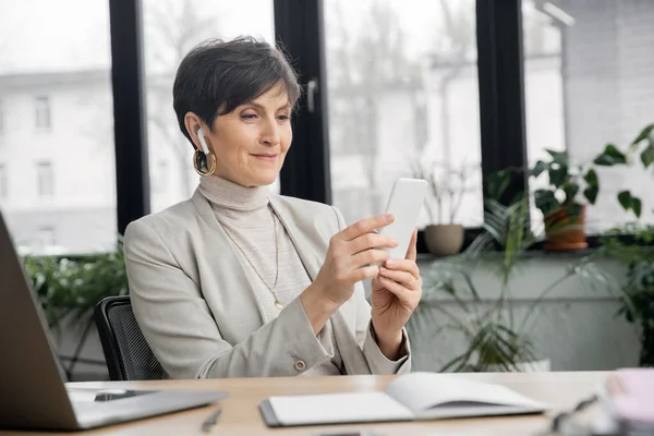 Pleased mature businesswoman in earbud browsing internet on smartphone near laptop in office — Stock Photo