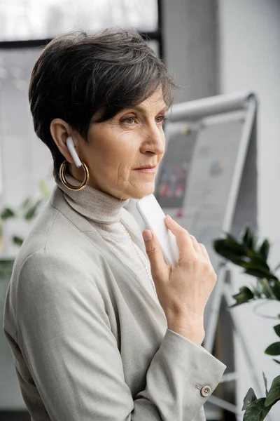 Pensive middle ages businesswoman listening music in earphones and looking away in office — Stock Photo