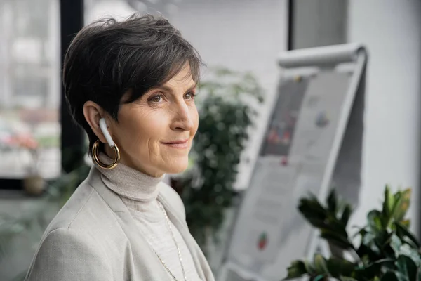 Positive and dreamy mature businesswoman listening music in earbud and looking away in office — Stock Photo