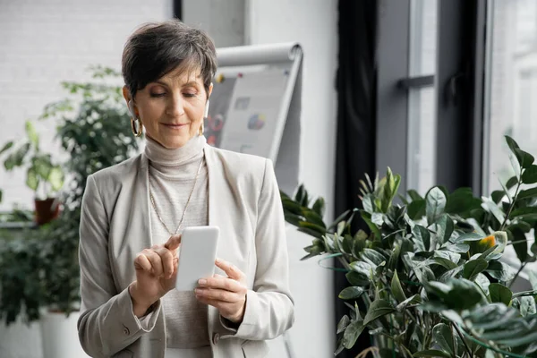 Successful middle aged businesswoman in earphones networking on smartphone in modern office — Stock Photo