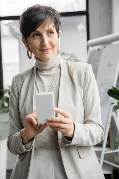 Smiling middle aged businesswoman in earphones holding smartphone and looking away in office — Stock Photo