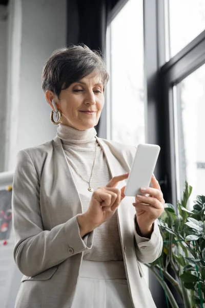 Mature woman, corporate manager listening music in earbud and networking on smartphone in office — Stock Photo