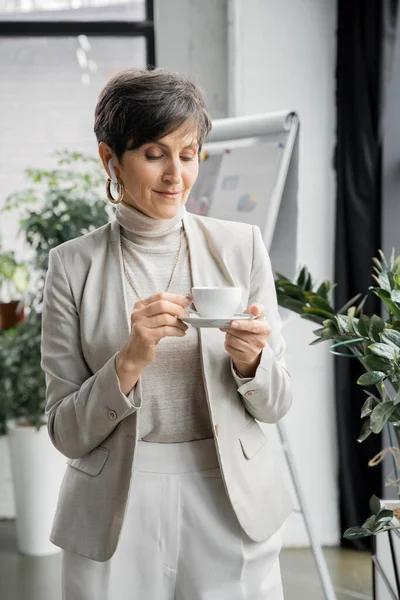 Femme d'affaires mature tenant une tasse de café et écoutant de la musique dans un écouteur dans un bureau moderne, plaisir — Photo de stock