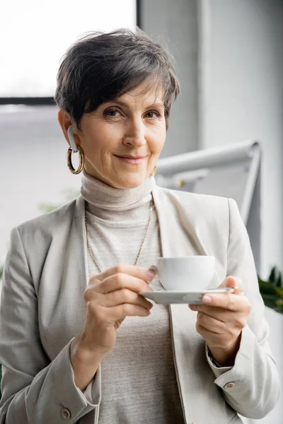 Portrait of stylish mature business woman in office, earphone, coffee cup, smiling at camera — Stock Photo