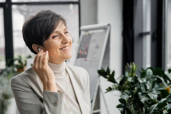 Overjoyed middle aged businesswoman adjusting earphone while listening music in office, enjoyment — Stock Photo