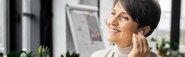 Pleased middle aged entrepreneur adjusting earbud and listening music in office, banner — Stock Photo