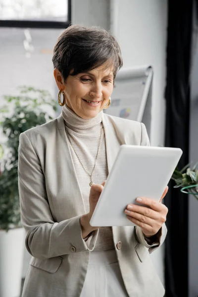 Sonriente y elegante mujer de negocios de mediana edad utilizando tableta digital en la oficina contemporánea - foto de stock