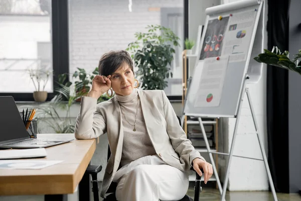 Femme d'affaires mature réfléchie sur le lieu de travail près d'un ordinateur portable et tableau à feuilles mobiles avec des graphiques, résolution de problèmes — Photo de stock