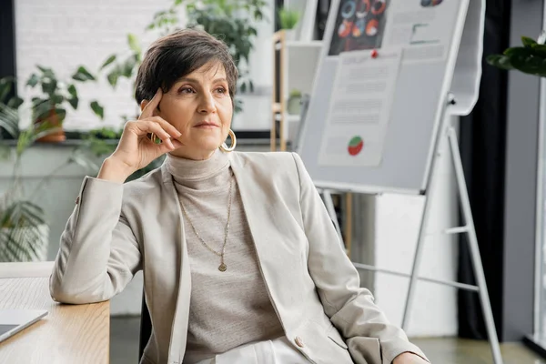 Coûteuse femme d'affaires d'âge moyen regardant loin près flou tableau à feuilles mobiles au bureau, pensée créative — Photo de stock