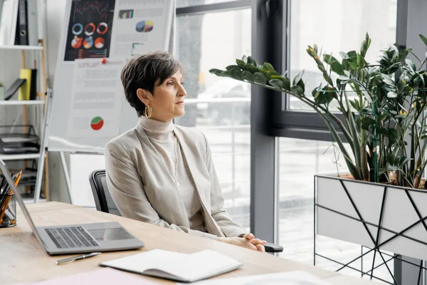 Mujer emprendedora reflexiva mirando hacia otro lado cerca del ordenador portátil y rotafolio con gráficos en la oficina moderna - foto de stock