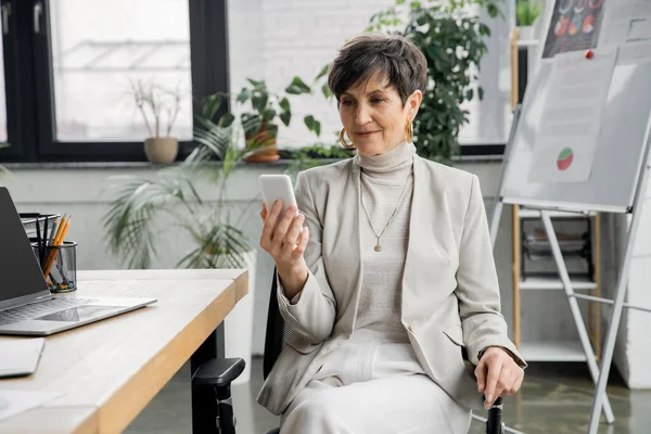 Positive Geschäftsfrau mittleren Alters mit Smartphone in der Nähe von Laptop und Flipchart auf verschwommenem Hintergrund — Stockfoto
