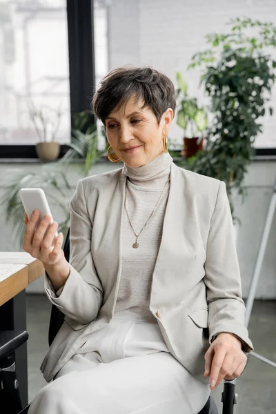 Lächelnde Geschäftsfrau mittleren Alters vernetzt sich am Arbeitsplatz im modernen Büro mit dem Smartphone — Stockfoto