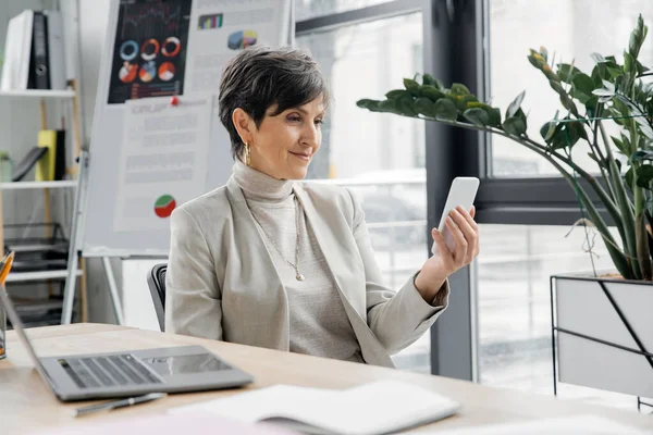 Mujer de negocios de mediana edad con teléfono inteligente cerca del ordenador portátil y rotafolio borroso con gráficos en la oficina - foto de stock
