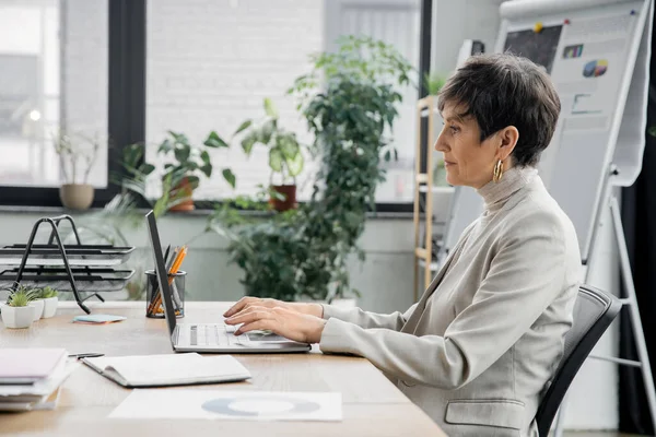 Вид збоку зрілої бізнес-леді, що друкує на ноутбуці поруч із записником та документами в сучасному офісі — стокове фото
