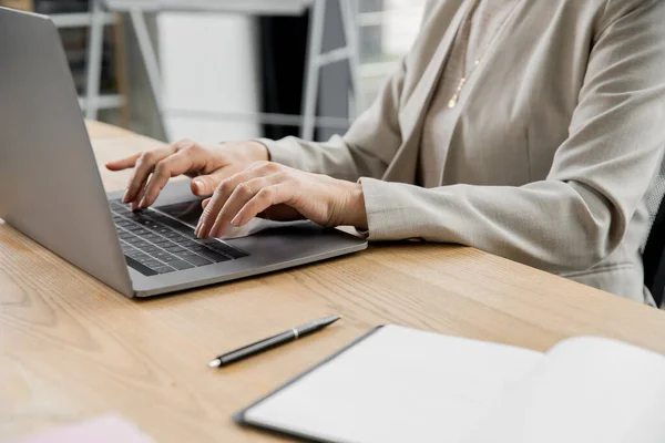 Частковий вид бізнес-леді, що друкує на ноутбуці поруч з ручкою і блокнотом на робочому столі в офісі — стокове фото