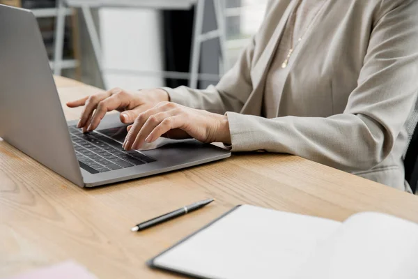 Partial view of businesswoman typing on laptop near pen and notebook on work desk in office — Stock Photo
