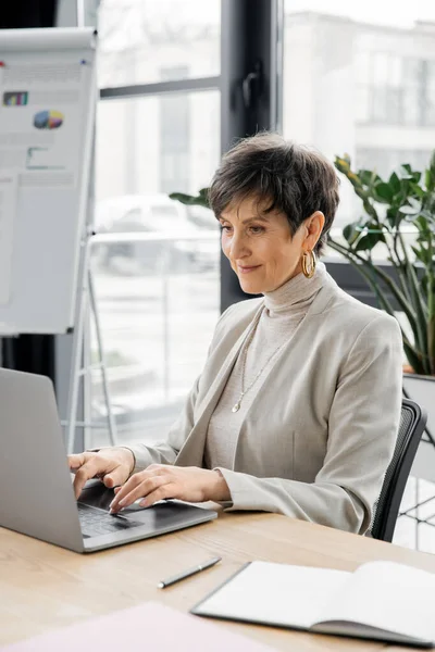 Experienced middle aged businesswoman typing on laptop near notebook at workplace in office — Stock Photo