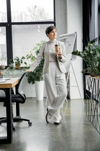 Повна довжина стильної бізнес-леді середнього віку з кавою, щоб піти стояти в сучасному офісі — стокове фото