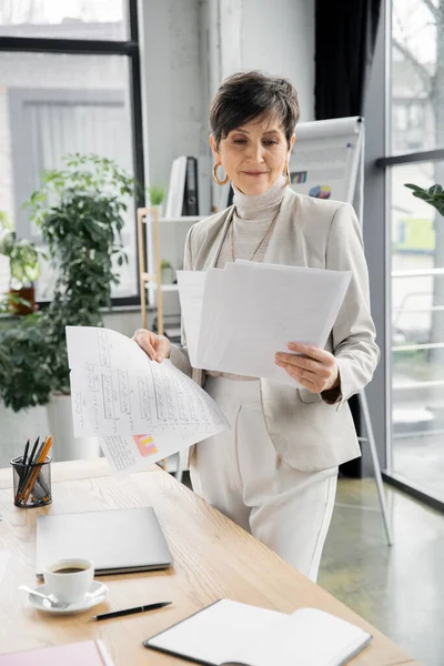 Middle aged businesswoman looking at documents near coffee cup and laptop at work place in office — Stock Photo