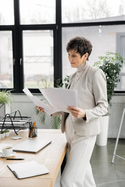 Confident middle aged businesswoman working with documents near laptop and coffee cup on work desk — Stock Photo