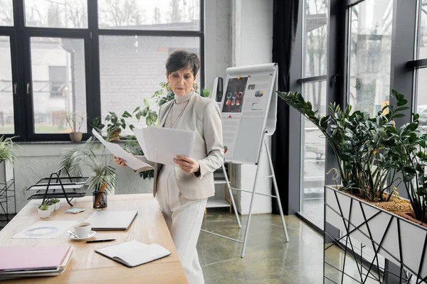 Stylish middle aged businesswoman with papers near coffee cup, laptop and notebook on work desk — Stock Photo
