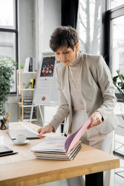 Modern middle aged businesswoman looking at documents near coffee cup in office, paperwork — Stock Photo